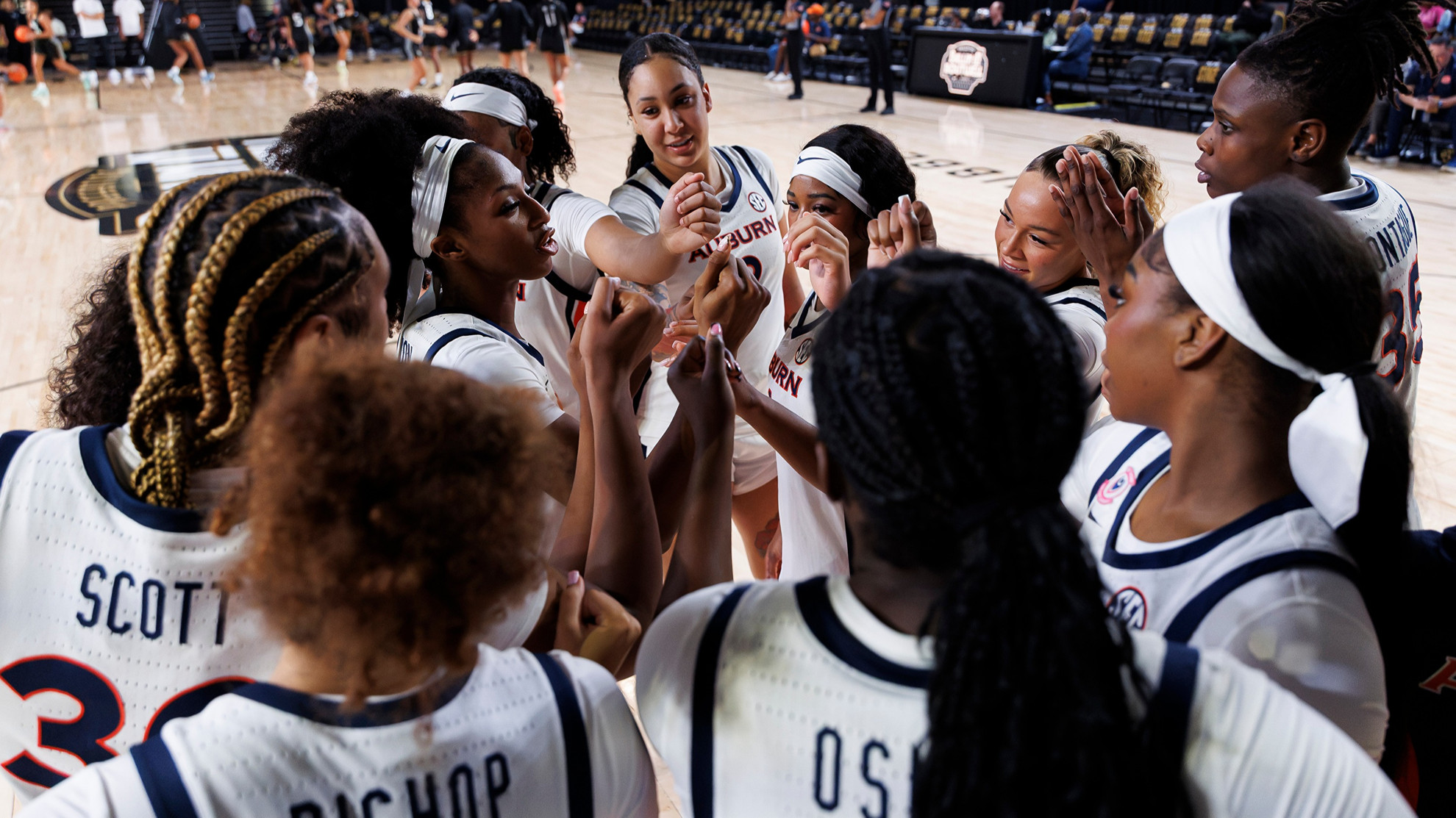 A group of basketball players in white jerseys huddle together on a court, with one player speaking to the team.