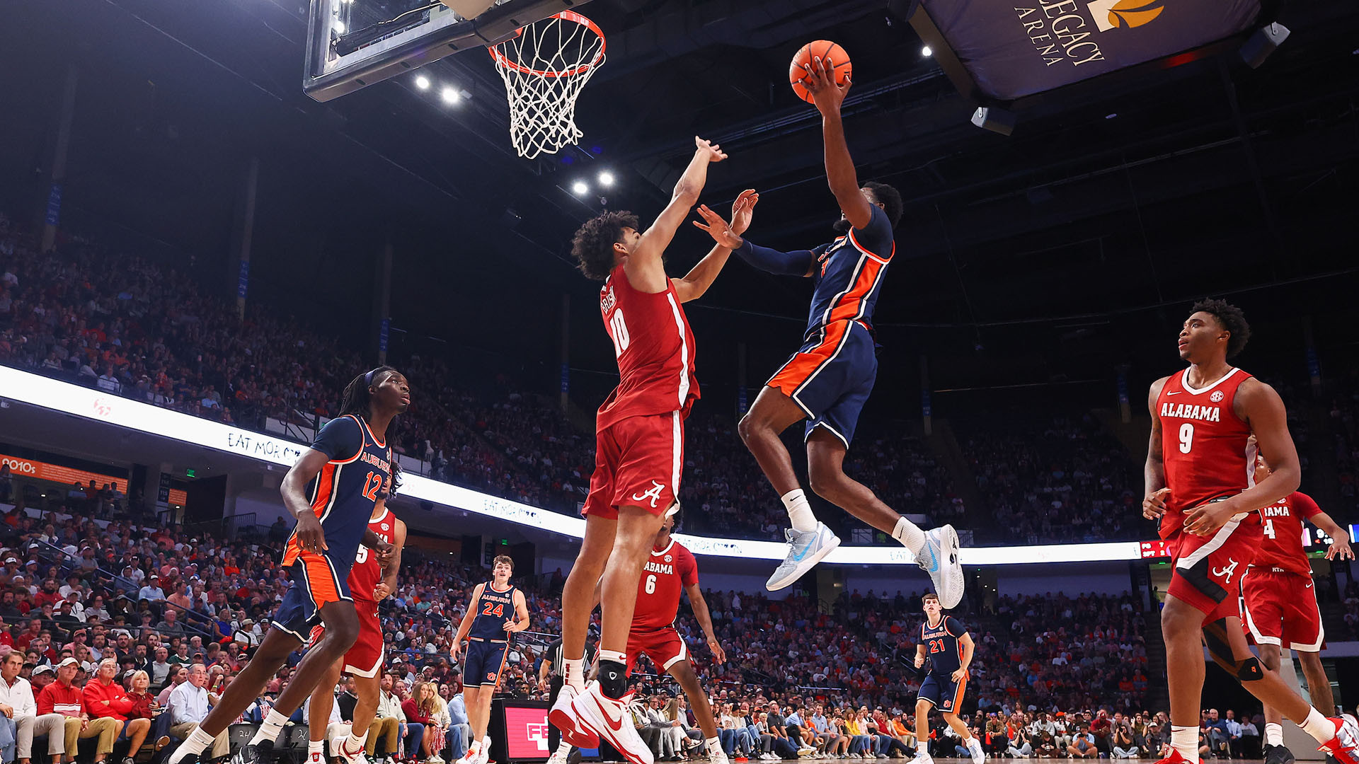 A basketball player in a blue and orange uniform leaps to shoot the ball while being defended by an opponent in a red uniform, with other players and a packed crowd visible in the background.