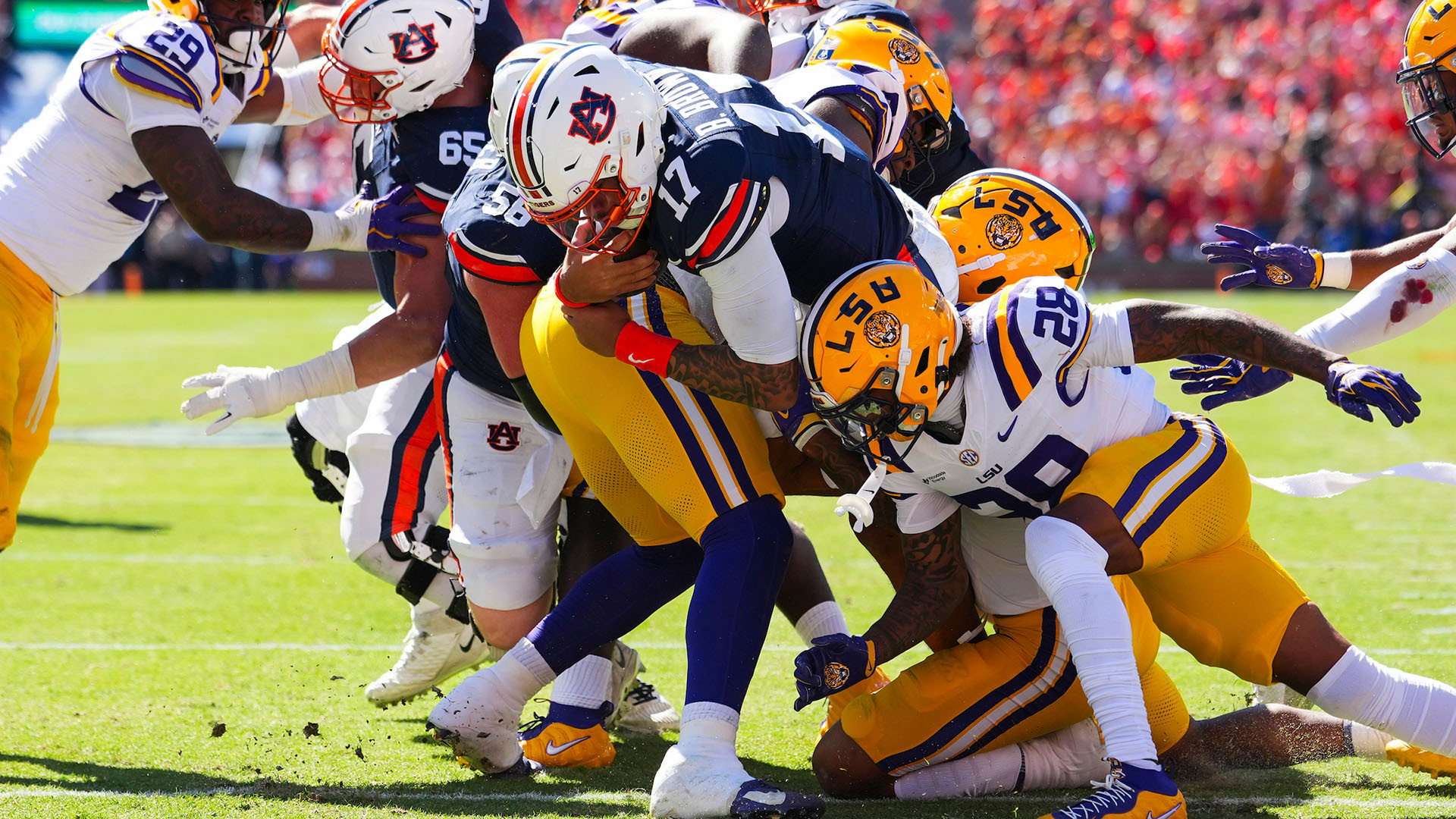Football players in yellow and purple uniforms engage in a tackle during a game on a sunlit field with a packed stadium in the background.